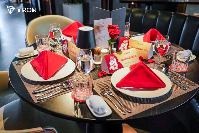 A circular table is set for four with red napkins folded into triangles, place cards featuring cartoon characters, a single red rose centerpiece, and a menu, all branded with the 'TRON' logo.