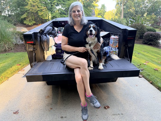 A smiling woman with gray hair sits in the back of a black pickup truck, accompanied by a happy Bernese Mountain Dog, with camping gear visible.