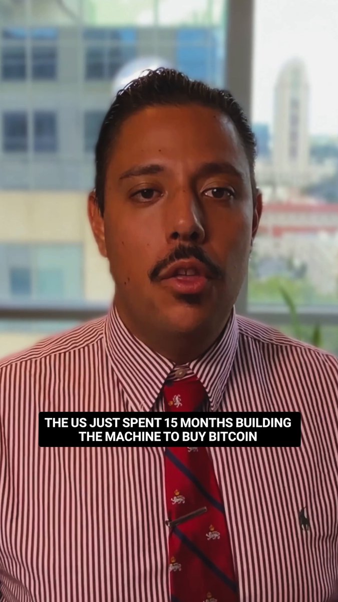 In the image, there's a man wearing a red striped shirt with a red tie around his neck. He has a mustache and is looking directly at the camera. The background of the photo shows a window overlooking an urban cityscape. There are also some text elements in the image that read "The US Just Spent 15 Months Building the Machine To Buy Bitcoin".