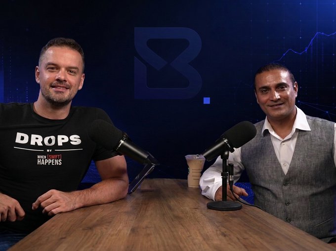 Two men sit at a wooden table with microphones in front of a blue background featuring a large 'B' logo and line graph; the man on the left wears a black 'DROPS' t-shirt.