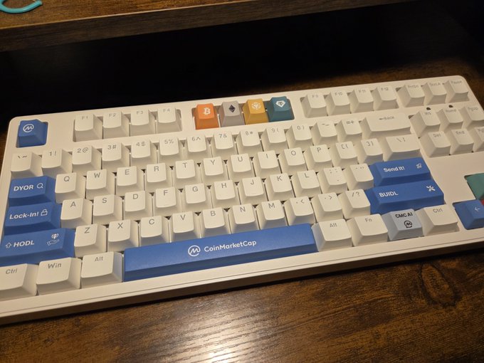 A white mechanical keyboard with blue accent keys labeled 'DVOR', 'BUIDL', 'HODL', and 'Lock-In!', featuring a CoinMarketCap logo on the spacebar and cryptocurrency-themed custom keycaps.