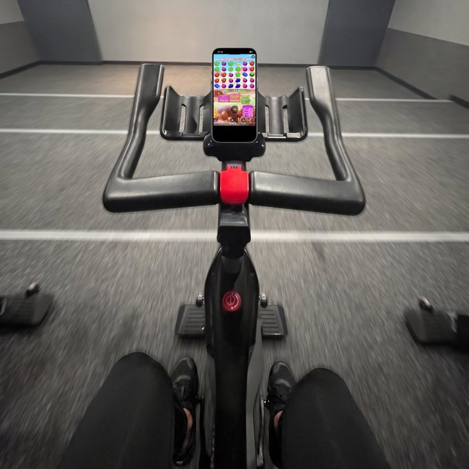 A first-person view of someone on a stationary bike with a smartphone displaying a home screen on the handlebars.