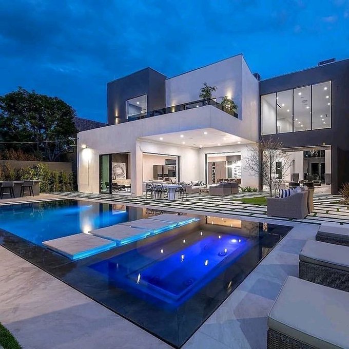 A modern luxury home at dusk featuring a swimming pool and spa with illuminated blue lighting, surrounded by a patio with lounge seating.