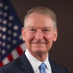 A portrait of SEC Chair Paul Atkins, smiling in a dark suit and blue tie, with an American flag visible behind him. The image accompanies news of his statement on the SEC's Regulation Crypto Assets proposal, positioning him as the official making this announcement.