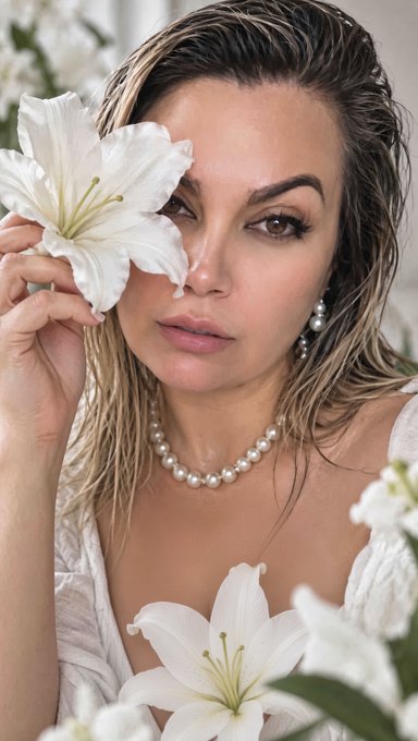 A woman with wet hair holds a white lily over one eye, wearing pearl jewelry including a necklace and earrings, with additional lilies in the foreground.