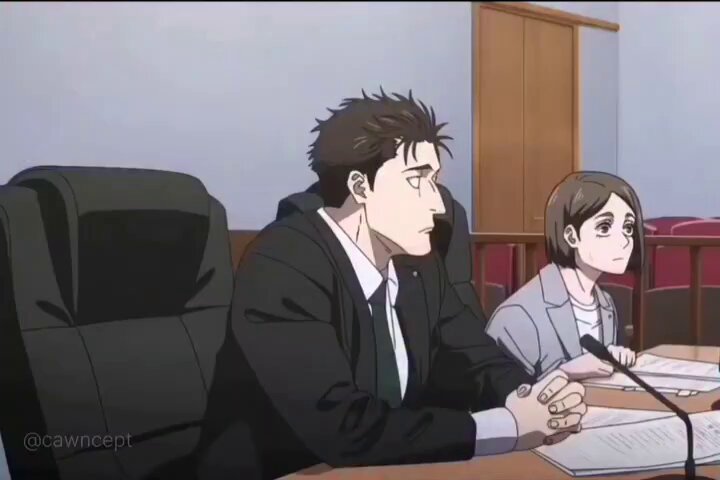 The image shows a man sitting at a desk with a woman behind him, both looking at something off camera. The scene appears to be set in an office or courtroom setting, as there are chairs and desks visible around them. The focus of the image is on the two individuals engaged in conversation or observing something outside their immediate surroundings.