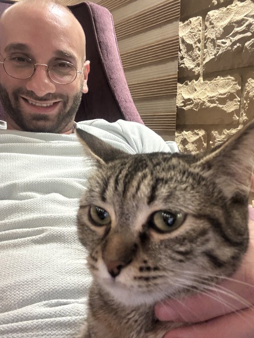 A bald man with glasses and a beard smiles warmly in a selfie while holding a brown tabby cat close to the camera.