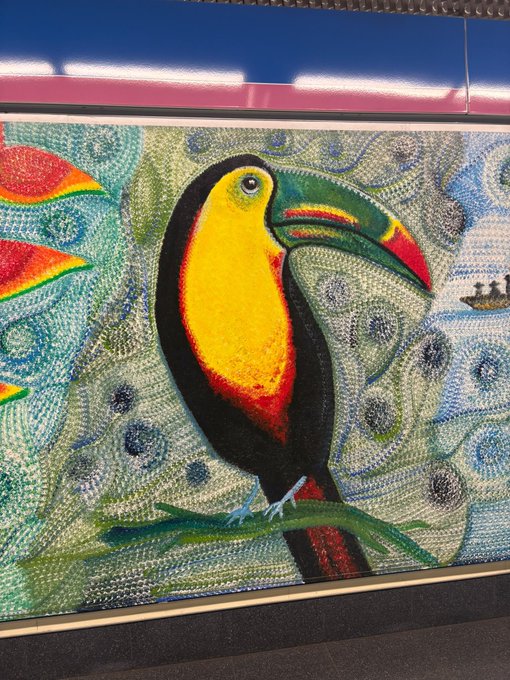 A photograph of a vibrant mural depicting a toucan perched on a branch, set against a patterned blue and green background with a row of bright lights visible at the top.