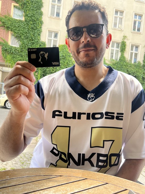 A man wearing sunglasses and a white and navy sports jersey with the text 'FURIOSO' and the number '17' holds up a black 'K/KART' Visa credit card outdoors.