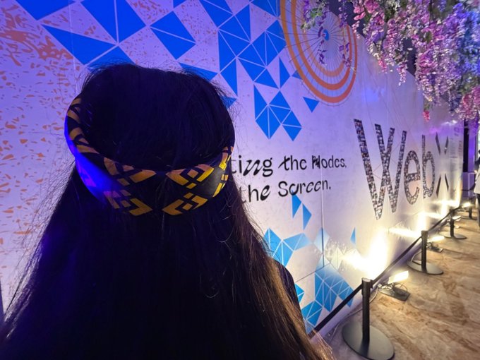 A person viewed from behind wearing a yellow and black patterned headband, standing in front of a wall with blue geometric designs and the text "wcv" and "izing the Nodes. the Screen."