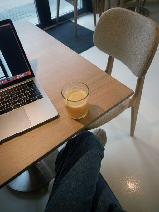 A first-person view of someone sitting at a wooden café table with a MacBook, a glass of orange juice, and their leg visible, facing a chair in a modern interior.