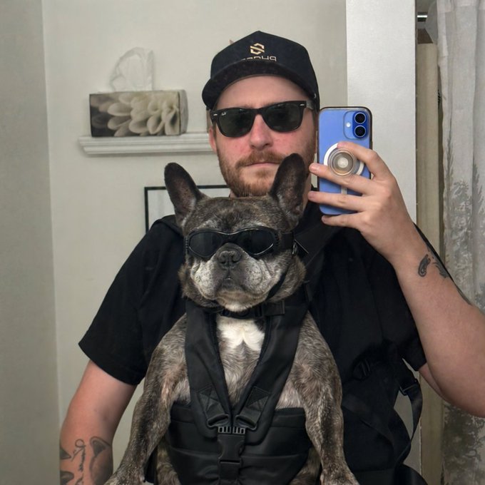 A mirror selfie showing a man in a black shirt, cap, and sunglasses holding a dog in a carrier, with the dog also wearing sunglasses.