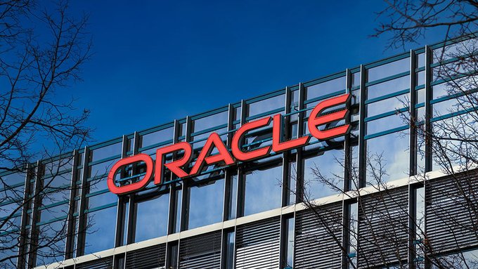 The red 'ORACLE' logo is displayed on the exterior of a modern glass-facade building against a clear blue sky.