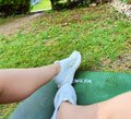 A person wearing light blue socks takes a photo of their feet propped up on a curb in a grassy area, matching the post's update about completing day 5 of the @0xAlphaGEMs Fitness Challenge with planks and core work.