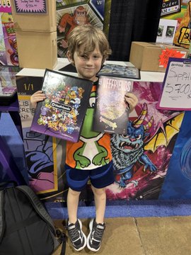 A young boy at what appears to be a comic convention, wearing a green and orange shirt and blue shorts, holding up two comic books for sale or display, with a 'Captain' sign showing '$3.00' visible in the background.