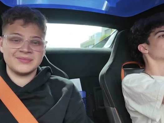 Two young men sit inside a car, with the passenger on the left wearing glasses and a black hoodie, both secured with orange seatbelts.
