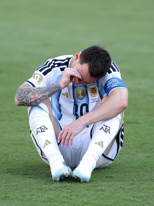 Lionel Messi, wearing Argentina's #10 jersey with the captain's armband, sits on the grass field wiping his eyes, appearing emotional after a match.