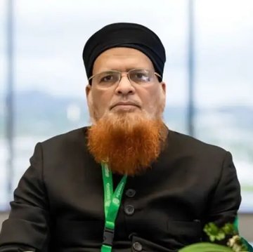 A bearded man wearing a black turban, glasses, and dark traditional attire with a green lanyard sits in what appears to be an official setting. The image likely depicts the Pakistani Islamic scholar referenced in the post who issued the fatwa declaring cryptocurrency purchases impermissible under Sharia law.
