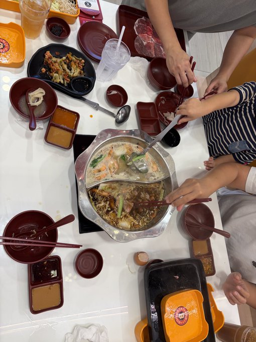 A group of people are sharing a meal at a restaurant, gathered around a divided hot pot containing a milky broth and a darker broth with vegetables, surrounded by small bowls and plates of food on a white table.