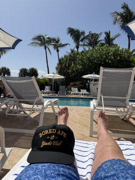 A relaxing poolside view from a lounge chair, showing bare feet resting on a towel with a black "BORED APE Yacht Club" cap placed nearby, overlooking palm trees and a resort swimming pool.