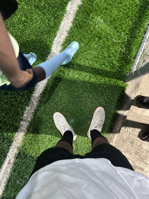 A first-person view shows two people standing on bright green artificial turf near a sideline, with one person wearing white sneakers and another wearing light blue shoes.