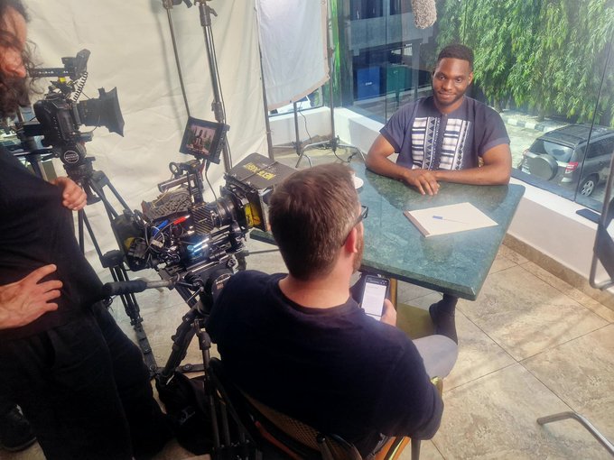 A film crew conducts an in-person interview with a young entrepreneur at a marble table outside a building, with a camera and lighting equipment visible. The setup reflects the context that Stripe went the extra mile—traveling over 10,000 km to Nigeria—to film an interview with a solo founder about stablecoins and building globally from Nigeria.