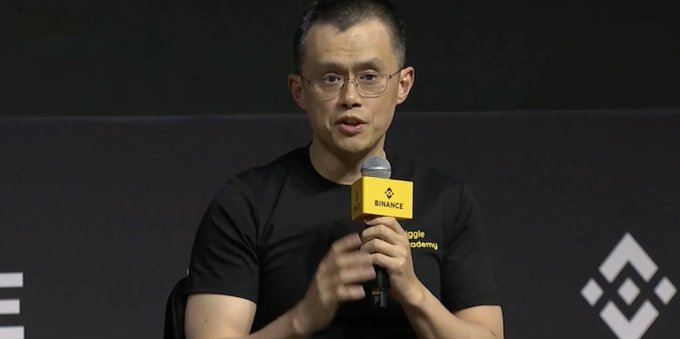 A man wearing glasses and a black Binance t-shirt speaking into a Binance-branded microphone at what appears to be a conference or panel discussion.