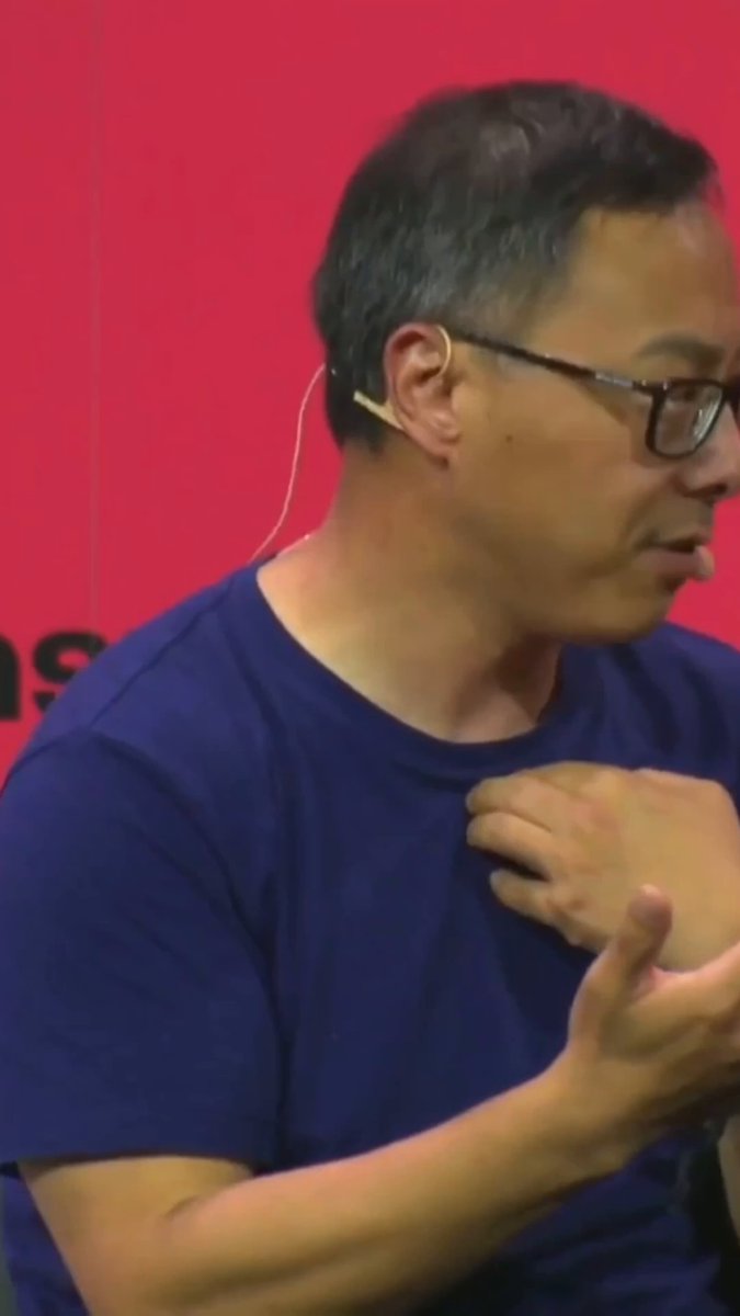 The image shows an older man wearing glasses and a blue shirt with his arm extended outwards. The background features a red wall that contrasts with the man's attire. There are no other people or objects visible in the image, making the focus on the man alone.