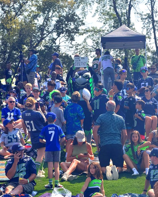 A crowd of Seattle Seahawks fans gathered outdoors to watch a game, with the mascot Blitz holding a sign reading "HAVE YOU SEEN JIMOTHY?" alongside a fan in a "METCALF 14" jersey. The scene ties into the post about "$Jimothy on @solana" by depicting a literal "Where's Waldo"-style hunt for "Jimothy" at a public fan event, playfully blending sports culture with crypto meme culture.