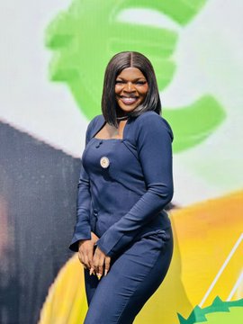 A smiling woman with shoulder-length black hair wearing a navy blue dress and a silver pendant necklace, posing in front of a green and yellow backdrop.