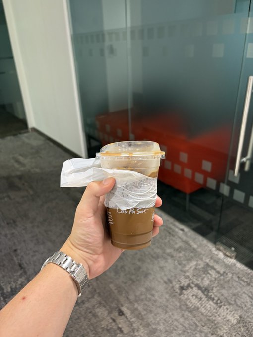 A hand wearing a silver watch holds an iced coffee in a plastic cup labeled "Cold Pla Sat," with a tissue wrapped around it for grip, set against an office backdrop with frosted glass and orange seating. The image of grabbing coffee suggests a casual meeting or networking moment, tying into the post about a productive call and plans for a Singapore event with @ImperialPerps and @clashofperps.