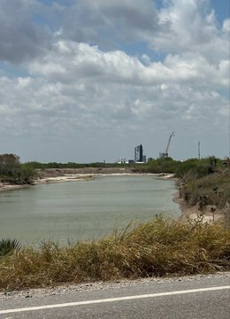 A landscape view of a greenish body of water with sandy shores, surrounded by vegetation, with industrial buildings visible in the distant background under a cloudy sky.