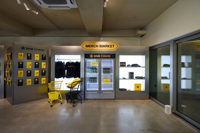 An indoor 'MERCH MARKET' pop-up shop displaying BNB Chain branded merchandise, hoodies, a beverage cooler, and yellow shopping carts.