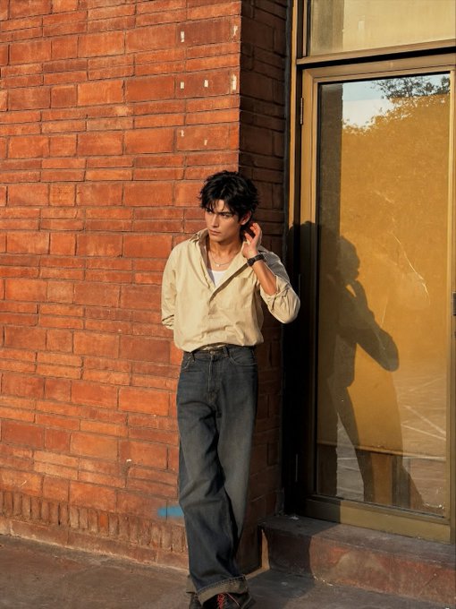 A young man with dark hair stands against a red brick wall next to a reflective window, wearing a cream-colored open shirt, white undershirt, blue jeans, and a watch, with his shadow visible in the glass.