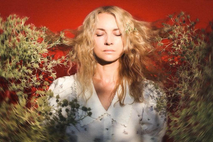 A woman with long blonde wavy hair and closed eyes, wearing a white floral blouse, photographed against a red background and surrounded by green foliage.