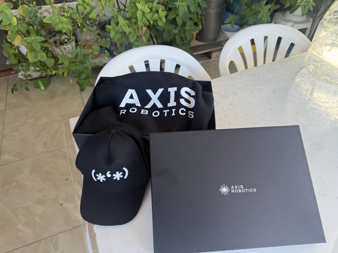 A black baseball cap with a white smiley face emoticon ( * * ) is displayed next to a black tote bag and black box, both branded with the 'AXIS ROBOTICS' logo in white text.