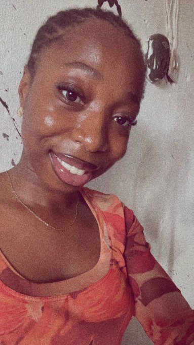 A woman with braided hair smiles warmly in a close-up selfie, wearing a coral-pink top against a white wall. The cheerful, bright-eyed expression complements the upbeat "good morning" greeting and encourages viewers to check out her pinned vlog and explore $RIVER.