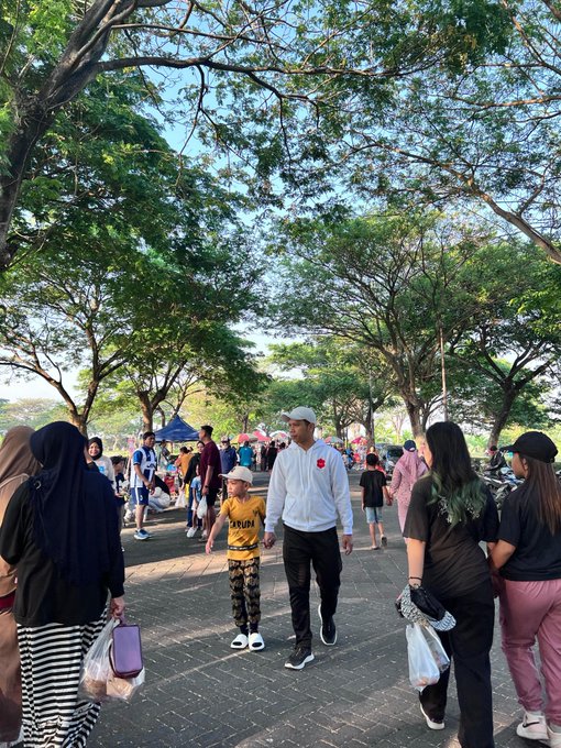A bustling outdoor pedestrian area shaded by large trees, with various people walking, including a man in a white hoodie and a boy in a yellow shirt.