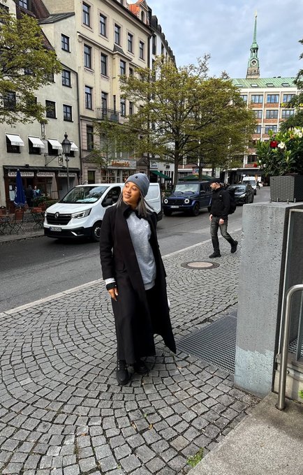A woman wearing a long black coat and gray beanie poses on a cobblestone street in a European city, with a clock tower and other pedestrians visible in the background.
