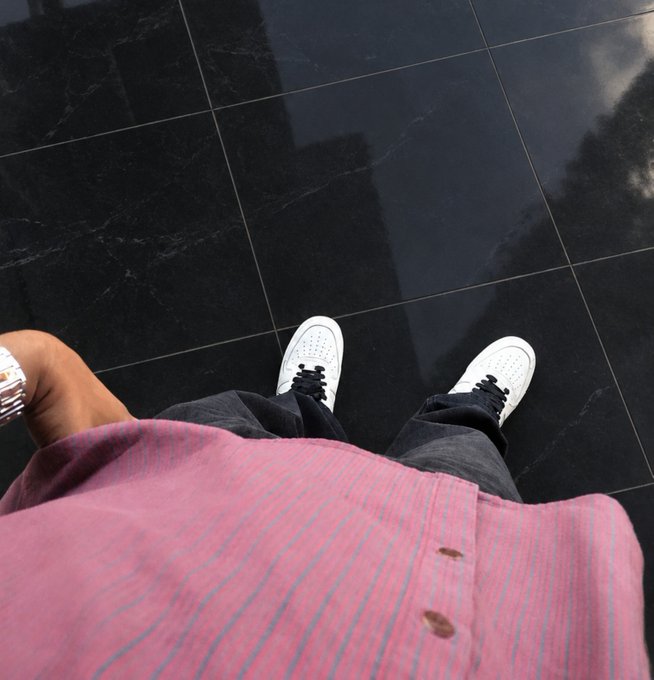 A downward-facing shot of a person wearing white sneakers, dark pants, and a pink striped shirt with a silver watch, standing on a glossy black tiled floor.