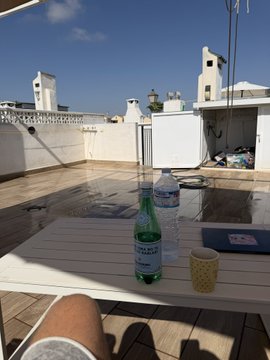 A rooftop terrace on a sunny day with a glass bottle of S.Pellegrino sparkling water, a plastic water bottle, and a small paper cup on a white table, overlooking white buildings under a clear blue sky.