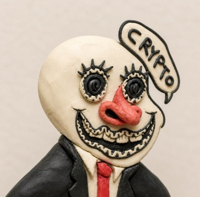 A close-up of a clay sculpture of a menacing clown figure with bulging eyes and a wide grin, wearing a black suit with a red tie, featuring a speech bubble that reads 'CRYPTO.'