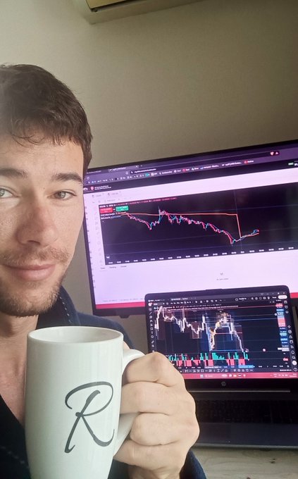 Selfie of a man holding a white mug with a cursive 'R' in front of a monitor and laptop displaying cryptocurrency price charts.