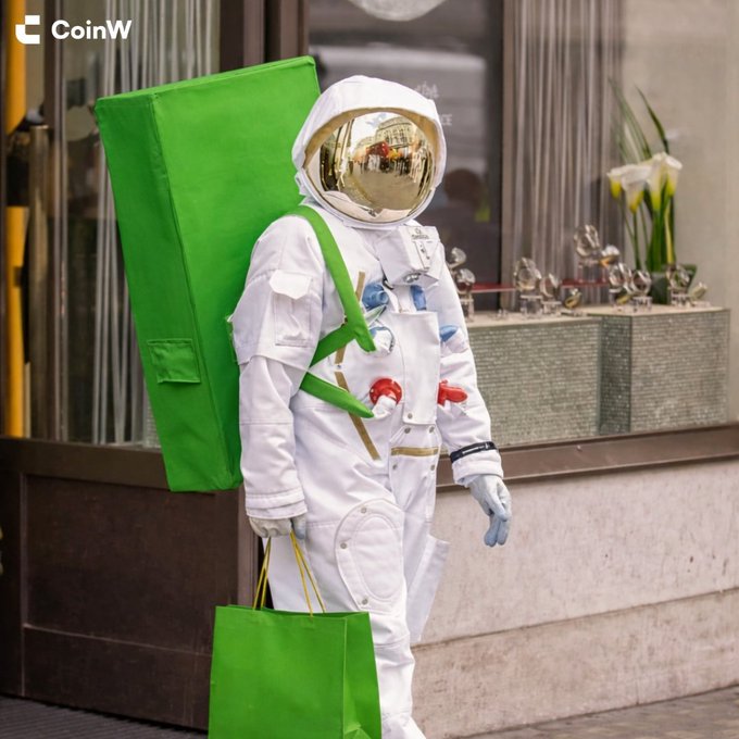 A person in a full white astronaut suit with a gold reflective visor stands on a city street, carrying a large green bag-shaped backpack and a smaller green shopping bag, with the "CoinW" logo visible in the top left corner. The image relates to the "GM" (Good Morning) post by creatively portraying an astronaut "arriving on Earth" to greet the crypto community.