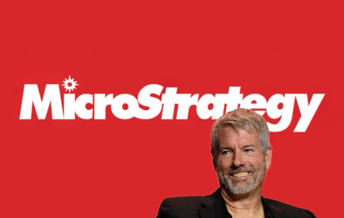 Michael Saylor, Executive Chairman of MicroStrategy, smiles in front of a red backdrop displaying the company's logo, visually connecting the tech executive to the ongoing debate over the firm's massive Bitcoin holdings and the billions in unrealized losses from market volatility.