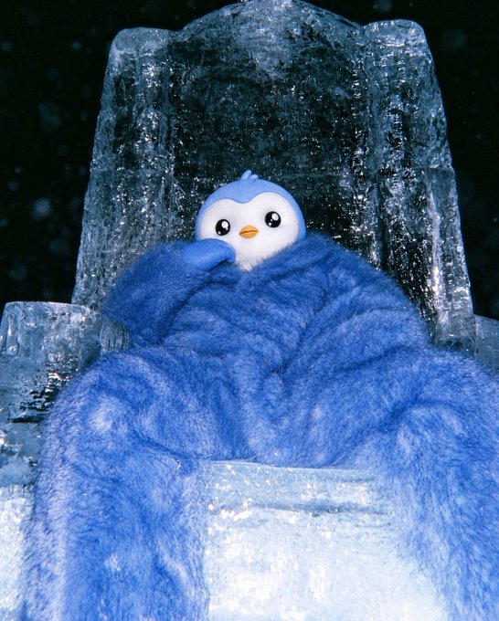 A small blue and white penguin plush toy wrapped in a fluffy blue blanket, sitting on an icy throne made of translucent blue ice against a dark background.