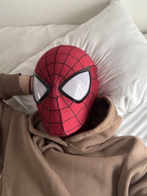A person wearing a brown hoodie and lying in bed, with their head covered by a Spider-Man mask featuring large white eye lenses, resting on a white pillow.