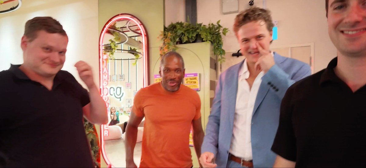 Four men smiling and gesturing in what appears to be a vlog setup, with one wearing an orange shirt and another in a blue blazer.