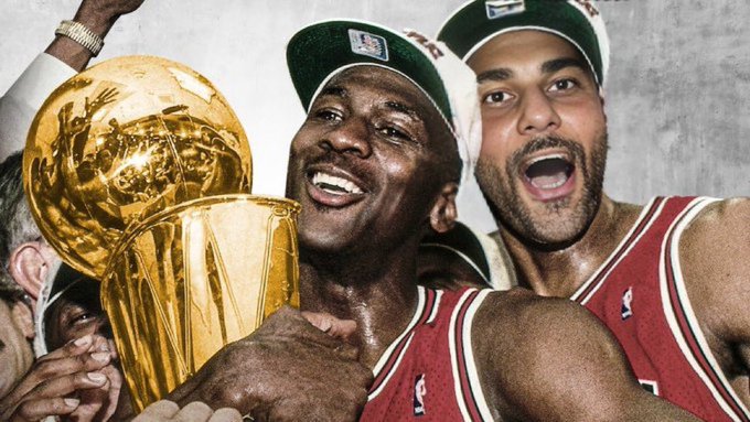 Michael Jordan and another player celebrate with the Larry O'Brien Championship Trophy while wearing Chicago Bulls championship hats and jerseys.