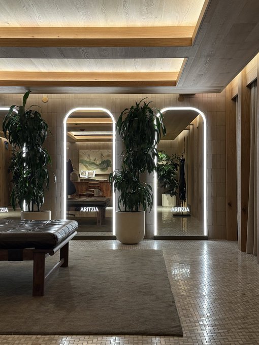 Interior shot of a stylish space featuring arched illuminated mirrors labeled "ARITZIA," flanked by large potted plants, with a cushioned bench on a tiled floor.